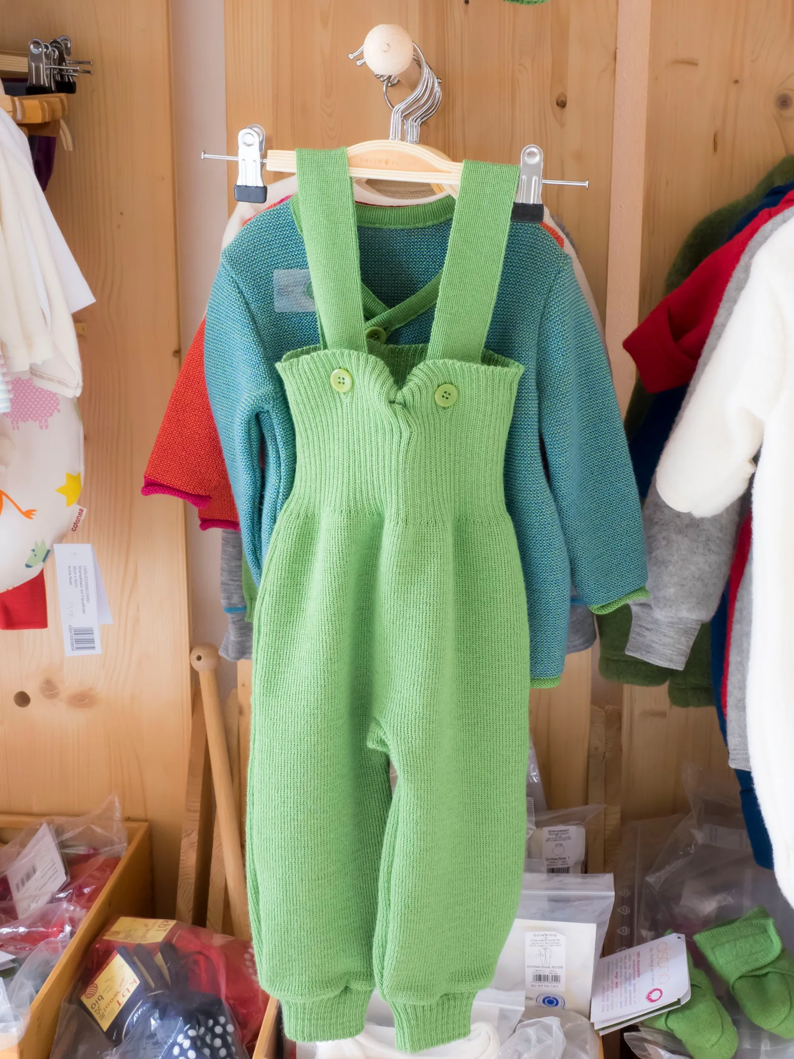 Grüne Strampelhose aus Wolle mit Trägern hängt vor einer Holzwand, dahinter Kinderpullover in Blau und Rot