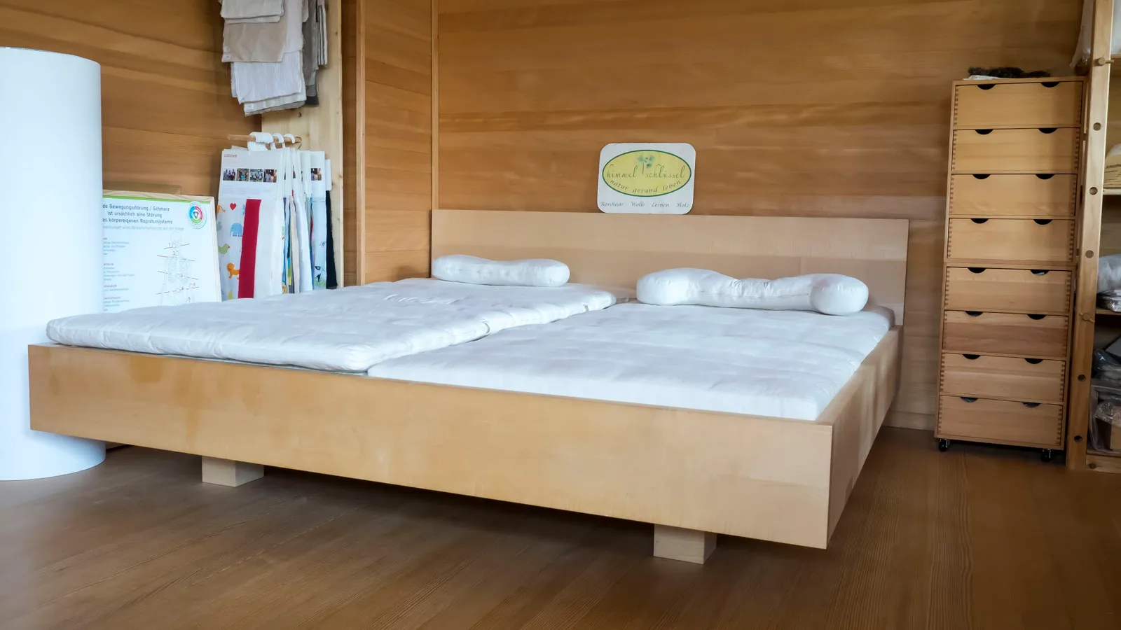 Naturbett aus hellem Massivholz mit zwei Matratzen und Nackenkissen, an der Holzwand darüber das Firmenschild mit dem Schriftzug himmelschlüssel