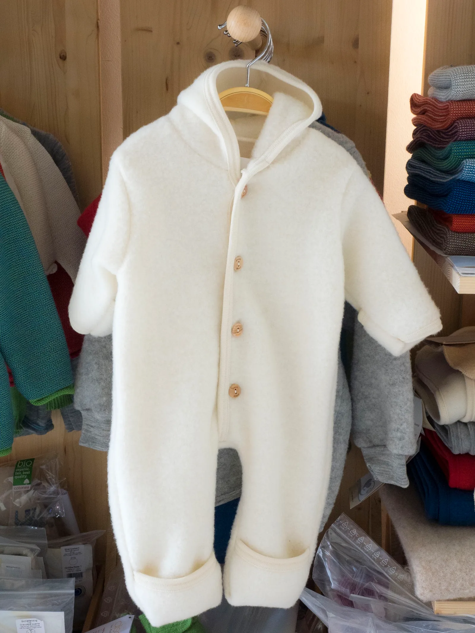 Cremeweißer Baby-Overall aus Wollfleece mit Kapuze und Holzknöpfen hängt vor einer Holzwand