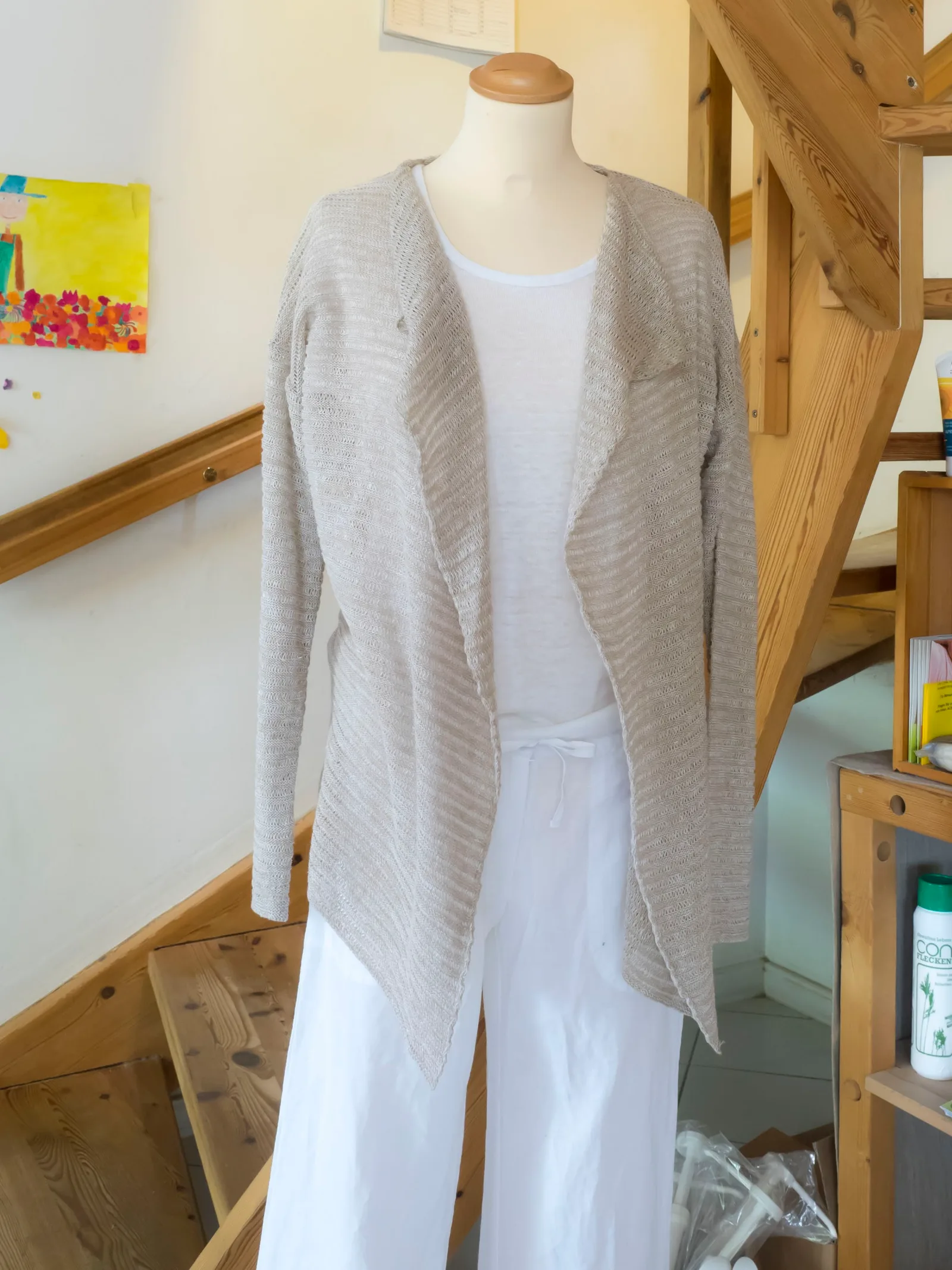 Beige Strickjacke aus Leinen über weißem Shirt und weißer Leinenhose auf einer Schneiderpuppe neben der Holztreppe