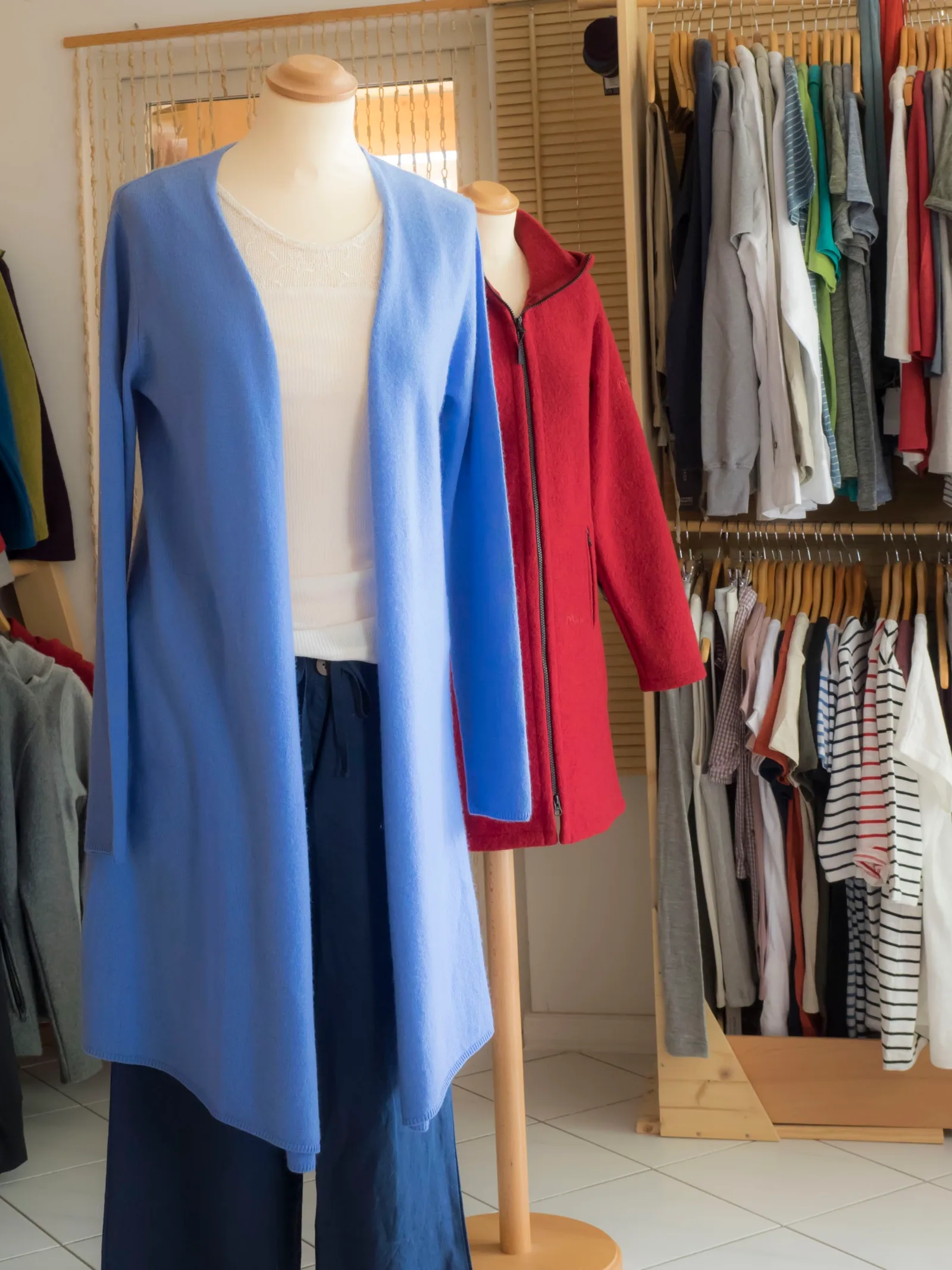 Verkaufsraum mit zwei Schneiderpuppen, einem hellblauen Longcardigan und einem roten Wollwalkmantel, dahinter volle Kleiderstangen