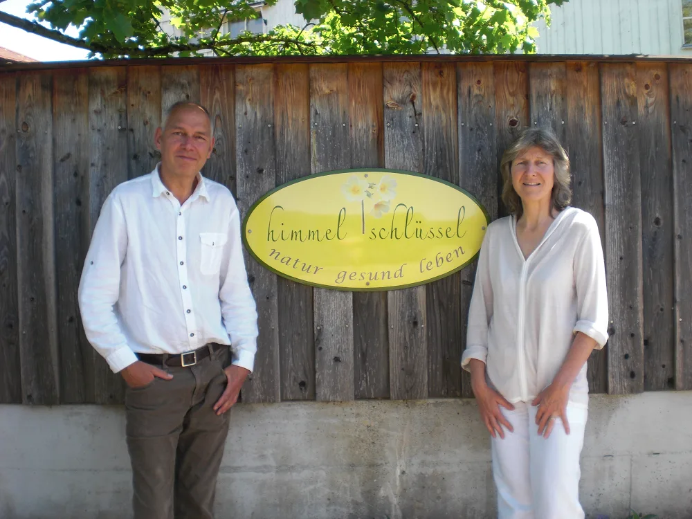 Helgard und Stefan Dilsky stehen links und rechts neben dem gelben ovalen Firmenschild an der Holzwand vor dem Haus